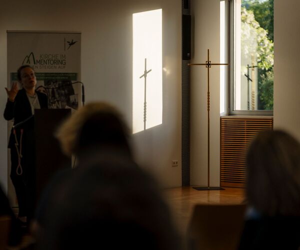 Kreuz aus Metall steht vor einem Fenster, der Schatten wird an eine weiße Wand geworfen; © Daniel Schwarz