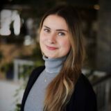 Louisa Harlow mit langem, dunkelblonden Haar und blauen, leicht geschminkten Augen, trägt einen grauen Rollkragenpulli und dunklen Blazer und lächelt leicht.