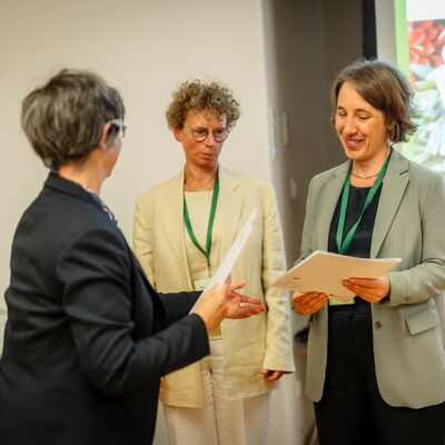 Drei Frauen: links mit dem Rücken zur Kamera, dunkler Blazer. in der Mitte hellgelber Blazer mit kurzen, lockigen Haaren. Rechts kurze, glatte Haare mit hellgrünem Blazer; © Daniel Schwarz