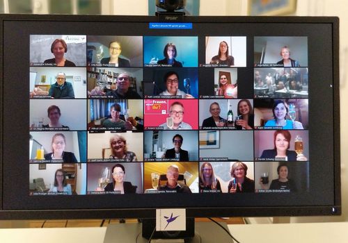 Foto eines Bildschirms, auf denen 25 Gesichter von Menschen zu sehen sind, die sich in einer Videokonferenz befinden. Viele heben Sektgläser hoch.
