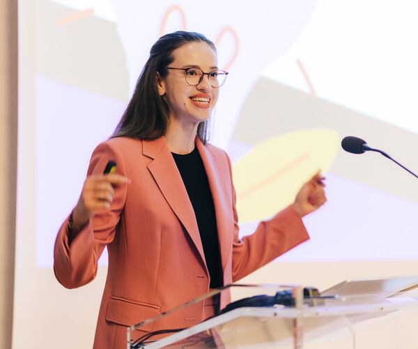Frau mit langen, dunklen Haaren, Brille, lachsfarbener Blazer steht vor Rednerpult mit Mikrophon, einladende Gestik; © Frank Herzmann