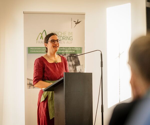 Frau in rotem Kleid mit braunen Haaren im Dutt steht vor einem Rednerpult mit Mikrophon und hält eine Ansprache; © Daniel Schwarz